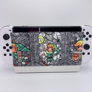 May include: A white Nintendo Switch console with a custom cover featuring a stained-glass style illustration of a character in green and yellow attire. The cover has a white base and a detailed, colorful design.