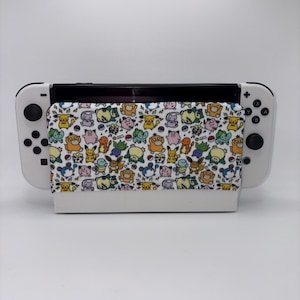 May include: A white Nintendo Switch console with a patterned cover featuring various cartoon characters in yellow, orange, blue, and green. The cover is wrapped around the console's screen, and the Joy-Cons are white.