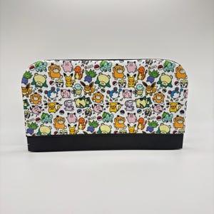 May include: A rectangular cosmetic bag with a black base and a white top covered in a colorful pattern of cartoon characters. The bag features a zipper closure and is designed for storing makeup and beauty products.