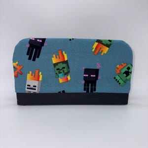 May include: A rectangular, blue fabric item with a black base, featuring a repeating pattern of pixelated video game characters in various colors. The characters include zombies, endermen, and creepers.