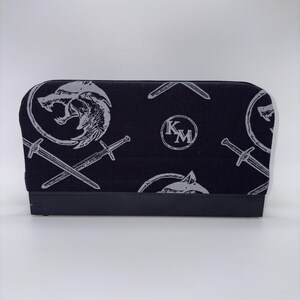 May include: Black and gray patterned case featuring a wolf head, crossed swords, and the letters "KM". The case has a black base and a rectangular shape. The design is repeated across the fabric.