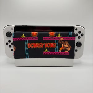 May include: A white handheld gaming console featuring a black fabric cover with a Donkey Kong print. The fabric displays the text DONKEY KONG, brown barrels, yellow bananas, blue ladders, and a brown gorilla character on a black background.