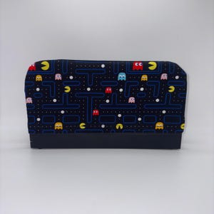 May include: A rectangular fabric cover featuring a black background with a repeating pattern of yellow Pac-Man characters, colorful ghosts in red, light blue, and pink, and blue maze lines with white dots. The base is a solid black rectangular block.