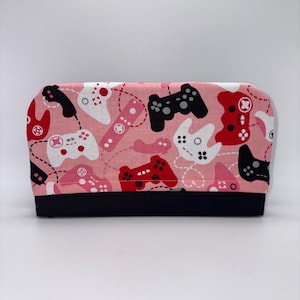 May include: A pink fabric item with a black base, patterned with white, black, and red video game controllers and remote controls. The controllers are connected by dotted lines. The item is likely a hat or head covering.