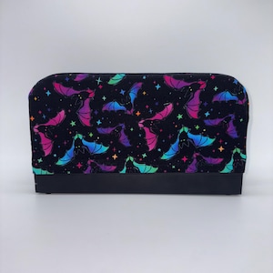 May include: A black rectangular cosmetic bag with a vibrant pattern of colorful bats and stars. The bats feature shades of pink, purple, blue, and green. The bag has a solid black base.
