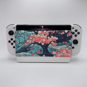 May include: A white Nintendo Switch console with a custom case featuring a Japanese-style design. The case displays a tree with pink blossoms against a dark blue background, with blue waves at the bottom.