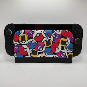 May include: A black handheld gaming console featuring a fabric cover with a repeating pattern of colorful spherical icons in red, blue, yellow, and purple. The cover displays various circular designs with white accents and black outlines.