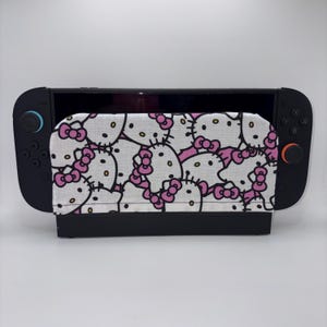 May include: A black Nintendo Switch console with a white and pink Hello Kitty patterned cover. The cover features a repeating pattern of Hello Kitty's face and pink bows. The console is on a white surface.