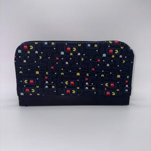 May include: A black zippered case with a Pac-Man video game pattern. The pattern features the game's maze, Pac-Man, ghosts, and power pellets in various colors. The case has a solid black base and a white background.