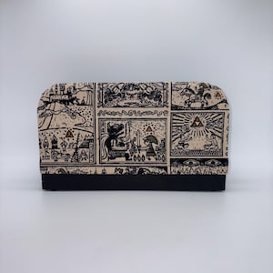 May include: A rectangular case with a black base and a beige top featuring a black illustrated design. The design includes various panels with figures, symbols, and scenes. The case is likely for storing items.
