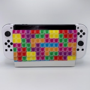 May include: A white Nintendo Switch console with a colorful, patterned cover. The cover features a repeating design of squares in various colors, including red, yellow, green, and purple. The console has black buttons and a black screen.