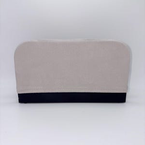 May include: A rectangular, light beige clutch with a black base. The clutch has a soft, textured appearance and a rounded top edge. The base is a solid, dark black color.