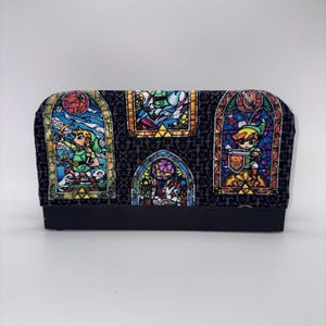 May include: A black rectangular box with a fabric top featuring stained-glass style illustrations of a video game character. The illustrations are in vibrant colors, set against a dark gray patterned background.