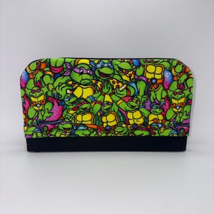 May include: A rectangular accessory featuring a vibrant, all-over print of the Teenage Mutant Ninja Turtles. The design showcases the turtles in various poses, with a black base. The color palette includes green, yellow, purple, and red.