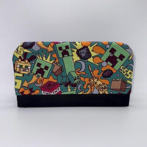 May include: A rectangular, fabric item with a black base and a colorful pattern featuring green Creeper characters, TNT blocks, and other elements from the popular video game Minecraft. The background is teal with yellow and orange accents.