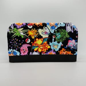 May include: A black fabric pouch featuring a colorful pattern of various cartoon characters and red and white ball graphics. The pouch has a solid black bottom and a curved top edge.