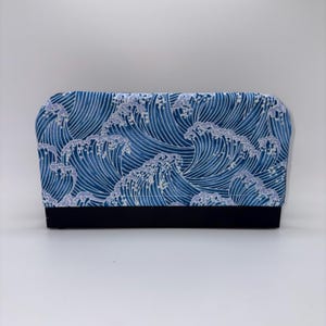May include: A rectangular zippered pouch featuring a repeating pattern of stylized blue and white ocean waves across the top section, contrasted by a solid black fabric base at the bottom. The design showcases curved lines and white foam details.
