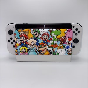 May include: A white Nintendo Switch console with a colorful fabric cover featuring various cartoon characters. The cover showcases characters like Mario, Luigi, and Yoshi against a blue background. The console's Joy-Con controllers are white with black buttons.