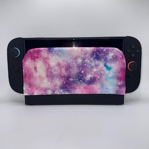 May include: A black handheld video game console featuring a fabric cover with a galaxy print in shades of pink, purple, and blue. The console has black controllers on each side, with a light blue joystick on the left and an orange ring on the right.