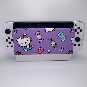 May include: A white Nintendo Switch console with a purple fabric cover featuring a Hello Kitty design and candy illustrations. The cover is secured to the console's screen, offering protection and a playful aesthetic. The design includes Hello Kitty, bows, and wrapped candies.