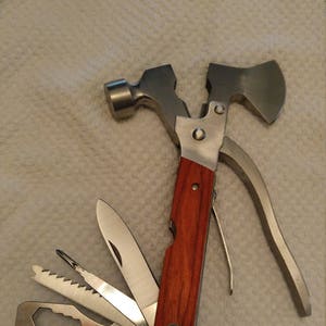 May include: A multi-tool with a hammer, axe, knife, saw, file, and other tools. The handle is wood-toned, and the metal components are silver. The tool is designed for various tasks and is compact for portability.