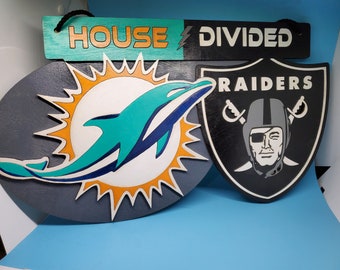 May include: Wooden sign with the text "HOUSE DIVIDED" and team logos. The sign features the Miami Dolphins logo and the Las Vegas Raiders logo. The Dolphins logo is a teal and white dolphin with an orange sun. The Raiders logo is a silver and black shield.