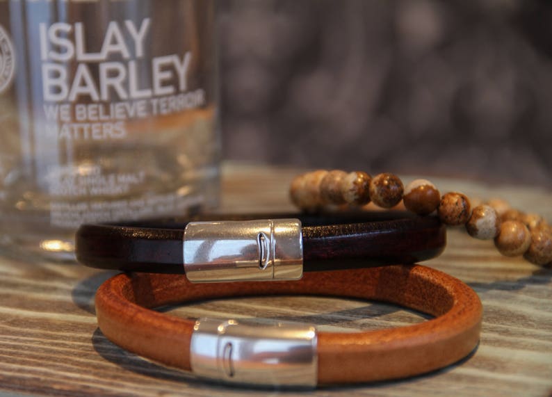 May include: Two leather bracelets with silver clasps. The top bracelet is dark brown and the bottom bracelet is light brown.