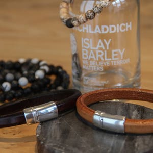 May include: Two leather bracelets with silver clasps. The bracelets are brown and tan. The bracelets are on a wooden surface.