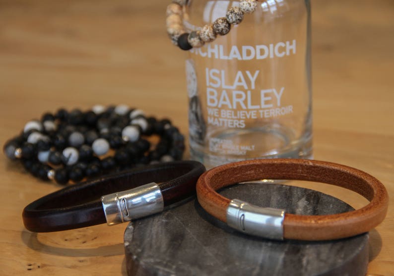 May include: Two leather bracelets with silver clasps. The bracelets are brown and tan. The bracelets are on a wooden surface.