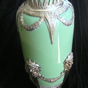 May include: A green ceramic vase with silver accents and ornate details. The vase has a wide mouth and a round base with three feet.
