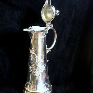 Puede incluir: Una jarra de plata con un diseño ornamentado y una tapa con bisagras. La jarra tiene un cuerpo alto y delgado con una base ancha y un pico en un lado. La tapa está decorada con un motivo floral. La jarra está hecha de metal y tiene un acabado pulido.