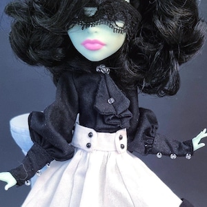 Peut inclure: Une poupée aux cheveux noirs bouclés, à la peau vert pâle et aux lèvres roses, vêtue d'une robe noire et blanche. La robe présente un corsage noir avec un col à volants et une jupe blanche. La poupée porte également un masque en dentelle noire.