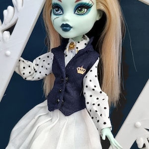 May include: A green-skinned doll with blonde hair wearing a white shirt with black polka dots, a navy blue vest with a gold crown pin, and a white skirt. The doll is posed against a white frame with ornate details.