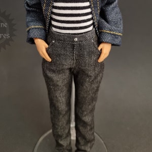 Puede incluir: Muñeco con chaqueta vaquera con costuras amarillas, camiseta a rayas blancas y negras y pantalón gris. El muñeco tiene pelo castaño y ojos azules. El muñeco está de pie con las manos en los bolsillos. Producto Sunshine Creatures.