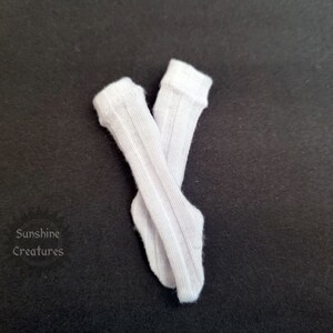 May include: Two white, ribbed knee-high socks, arranged in a cross against a dark backdrop. The socks are made of a soft material and feature a traditional design. The text "Sunshine Creatures" is visible in the lower left corner.