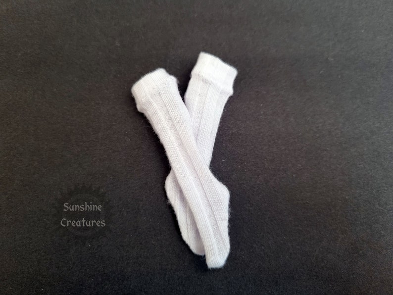 May include: Two white, ribbed knee-high socks, arranged in a cross against a dark backdrop. The socks are made of a soft material and feature a traditional design. The text "Sunshine Creatures" is visible in the lower left corner.