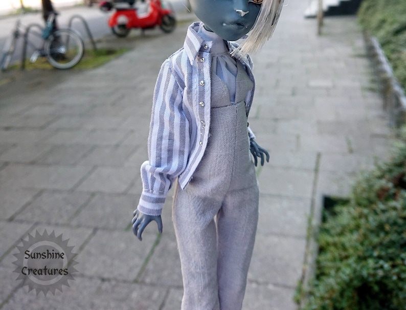 A Monster high doll wearing a light gray jumpsuit and a white and gray striped button-down shirt. The image is taken outdoors on a paved surface.