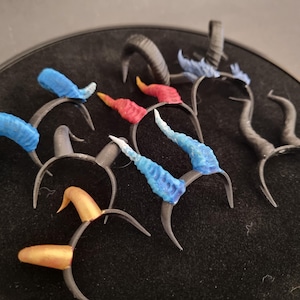 May include: A collection of fantasy-themed headbands with various horn designs. The horns are in shades of blue, red, gold, and black, attached to dark gray headbands. The horns vary in shape and texture, creating a unique assortment.