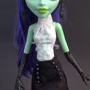 May include: A green-skinned doll with purple hair wearing a white and black outfit. The doll has black gloves with claw-like fingers.
