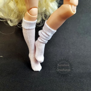 May include: A close-up of doll legs wearing white ribbed knee-high socks. One sock is pulled up, the other is scrunched down. The doll's legs are a light skin tone. The background is black. The text "Sunshine Creatures" is in the lower right corner.