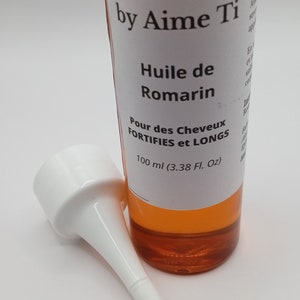 May include: A clear glass bottle with a white label that reads "Boucl'Ebene by Aime Ti Huile de Romarin Pour des Cheveux FORTIFIES et LONGS 100 ml (3.38 Fl. Oz)". The bottle contains a yellow liquid and has a white plastic applicator tip.