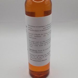May include: A clear glass bottle with a white label containing text in French. The bottle is filled with a light amber liquid. The label reads "Stimulante et tonique, le Romarin... antipelliculaire et stimulateur de peau. Associ&eacute; &agrave; l'huile de Colza, vos cheveux seront brillants d&egrave;s la premi&egrave;re application. En usage seul, ou alli&eacute; &agrave; d'autres huiles et/ou beurres, vous pouvez utiliser cette huile sur la peau et le corps. Ingr&eacute;dients: Huile de Colza, Feuilles de Romarin Mac&eacute;r&eacute;es. Fabriqu&eacute; et distribu&eacute; par Boucle d'Eb&egrave;ne Ti- 11 rue du petit pont Epinay-sous-Senart. Mail:contact@boucl-ebene.com"