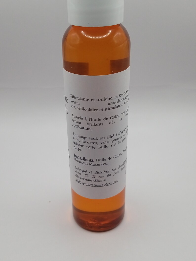 May include: A clear glass bottle with a white label containing text in French. The bottle is filled with a light amber liquid. The label reads "Stimulante et tonique, le Romarin... antipelliculaire et stimulateur de peau. Associ&eacute; &agrave; l'huile de Colza, vos cheveux seront brillants d&egrave;s la premi&egrave;re application. En usage seul, ou alli&eacute; &agrave; d'autres huiles et/ou beurres, vous pouvez utiliser cette huile sur la peau et le corps. Ingr&eacute;dients: Huile de Colza, Feuilles de Romarin Mac&eacute;r&eacute;es. Fabriqu&eacute; et distribu&eacute; par Boucle d'Eb&egrave;ne Ti- 11 rue du petit pont Epinay-sous-Senart. Mail:contact@boucl-ebene.com"