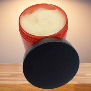 May include: An open, red jar filled with a creamy, off-white substance. The black lid is placed next to the jar. The product is on a wooden surface with a gradient background.