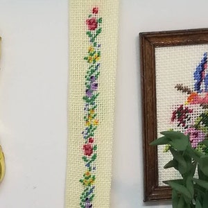 Puede incluir: Una pared de tela blanca con un gancho de metal dorado en la parte superior. La tela está bordada con una fila vertical de flores de colores.