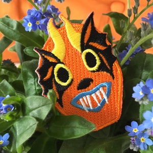 May include: Orange and black embroidered patch of a monster face with yellow horns, white teeth, and blue mouth. The patch is surrounded by green leaves and blue forget-me-not flowers.