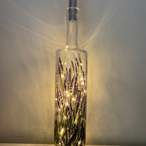 May include: A clear glass bottle with a cork stopper, decorated with a painted design of lavender flowers and a butterfly. The bottle is illuminated with warm white fairy lights.