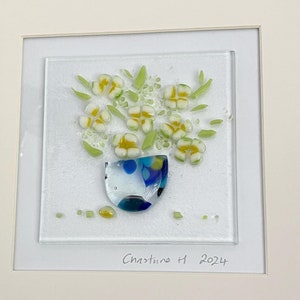 May include: A framed piece of art featuring a white vase with yellow flowers made of glass. The vase has a blue and green pattern. The art is signed "Christine H 2024".