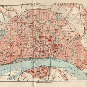 May include: A vintage map of Köln, Germany, featuring a detailed street layout in red and white, with the Rhine River running through the center. The map includes text in German and is printed on aged paper.