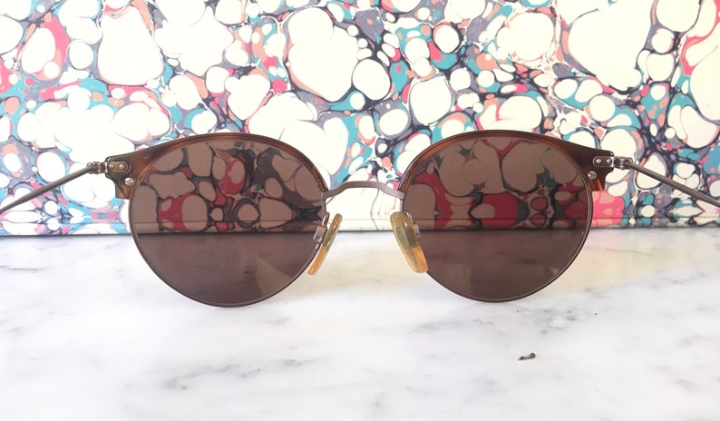 Puede incluir: Un par de gafas de sol redondas con montura marr&oacute;n y lentes marrones. Las gafas reflejan un patr&oacute;n de m&aacute;rmol.